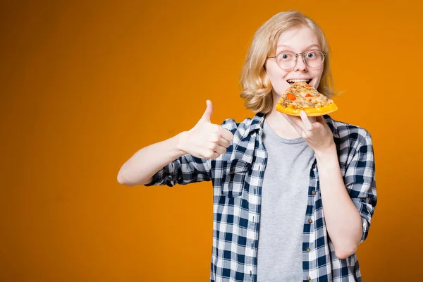 Pěkná blondýnka v brýlích jí plátek pizzy, usmívá se a ukazuje palec nahoru na oranžovém pozadí — Stock fotografie