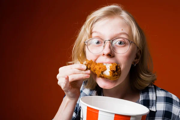 Nahaufnahme Porträt eines blonden Mädchens, das in ein Hühnerbein beißt und einen Eimer mit Hühnern in ihren Händen auf rotem Hintergrund hält — Stockfoto