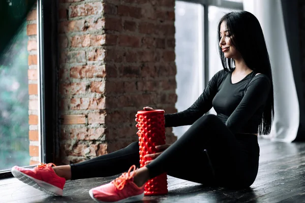 Sittande porträtt av en fitnesstränare i svart åtsittande kostym på golvet och med en massagecylinder för att stärka fasciamusklerna i gymmet — Stockfoto