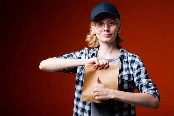 Μια ξανθιά κούριερ με μπλε καπέλο κρατά μια χάρτινη σακούλα και δείχνει τον αντίχειρα πάνω σε κόκκινο φόντο.. — Φωτογραφία Αρχείου