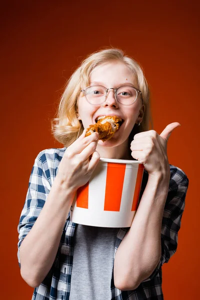 Vertikální fotografie blondýny v brýlích, která jí kuře a ukazuje jako na kameře na červeném pozadí. — Stock fotografie