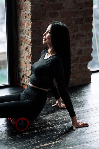 Vertikalt porträtt av en tjej i svart träningsoverall som lutar händerna på golvet och sitter med höfterna på en massagecylinder medan hon arbetar med fascian i de gluteala musklerna — Stockfoto