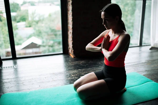 Vedere laterală a unei fete meditatoare antrenor yoga îngenunchind pe un covor sportiv în sala de sport vizavi de fereastră — Fotografie, imagine de stoc