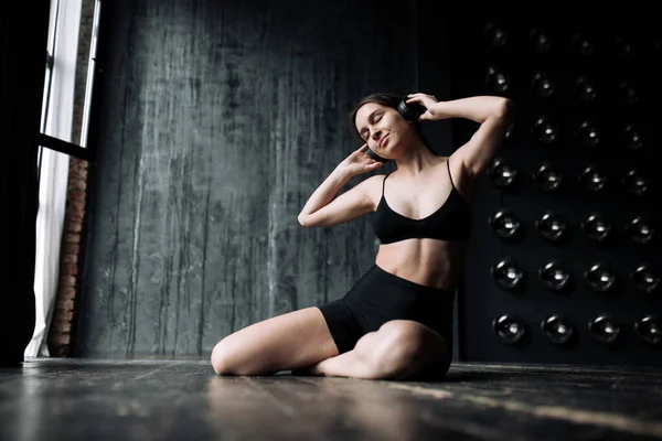 The girl is a yoga coach sitting in a lotus position and meditating while listening to music on headphones in the gym next to the panoramic windows.