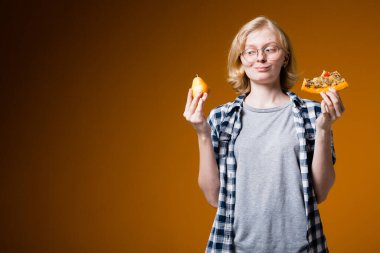 Gözlüklü sarışın kız, turuncu arka planda bir dilim pizzayı tutarken olgun bir armudu ısırıyor.