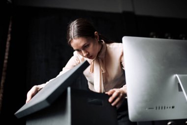 Ofisteki bir kara kutuda evrakları arayan bej gömlekli bir iş kızı portresi. Yakınlarda bir monitör var..