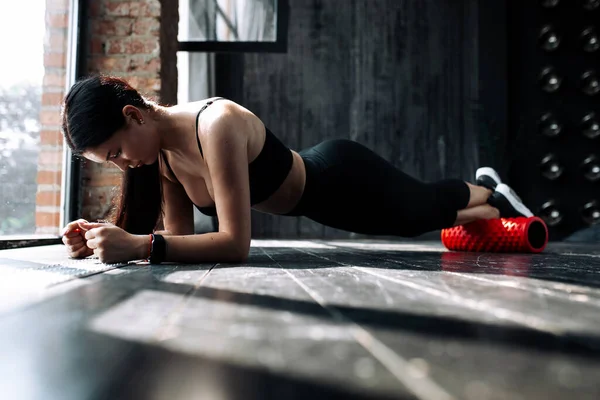 Una entrenadora de Pilates en ropa deportiva se sienta en un rollo rojo de masaje y se apoya en sus manos mientras trabaja en la fascia de los músculos de la espalda de las piernas . — Foto de Stock