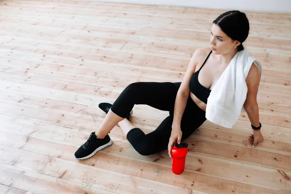 Vista superior de una entrenadora de pilates sosteniendo una coctelera roja con una toalla en los hombros sentada en el suelo con ropa negra ajustada —  Fotos de Stock