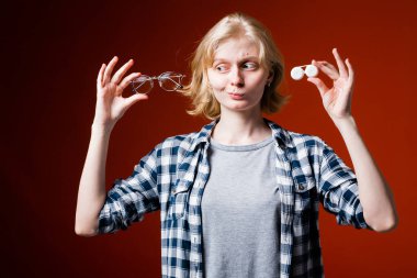 Gözleri iyi görmeyen sarışın kız elinde lensler ve gözlüklerle kırmızı arka planda ne seçeceğini bilemeyen bir kutu tutuyor.