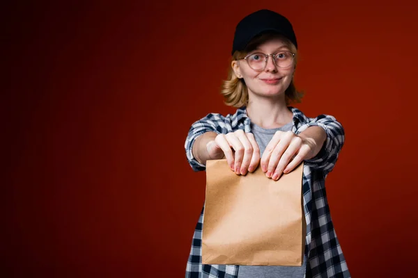 Γυναίκα ξανθιά υπάλληλος δίκτυο των εστιατορίων fast food στο πουκάμισο και το καπέλο κρατώντας ένα μαγειρεμένο φαγητό παραγγελία σε μια χάρτινη σακούλα. — Φωτογραφία Αρχείου