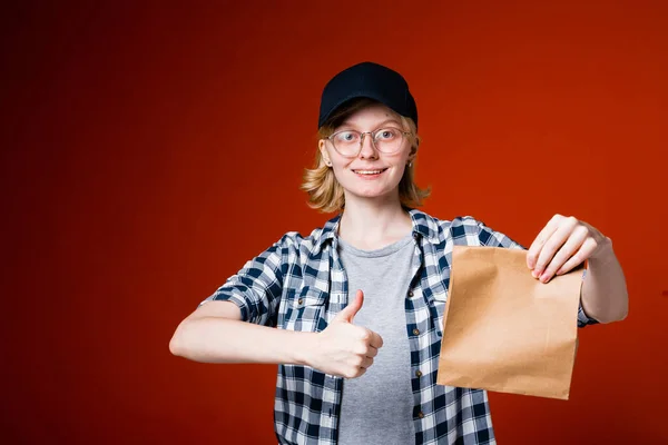 Κορίτσι courier σε ένα καπάκι κρατά μια χάρτινη τσάντα με φαγητό και δείχνει ένα παρόμοιο ως ένδειξη της γρήγορης και ποιοτικής παράδοσης — Φωτογραφία Αρχείου