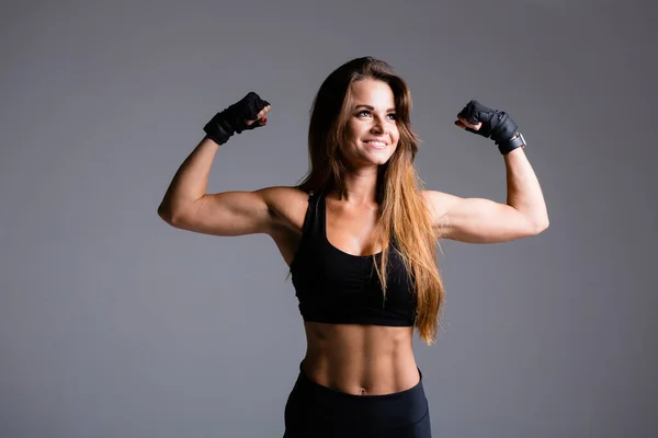 Gyönyörű bokszoló nő hosszú hajú mutatja kar izmok, bicepsz és tricepsz szürke alapon kesztyűben fekete sport öltöny — Stock Fotó