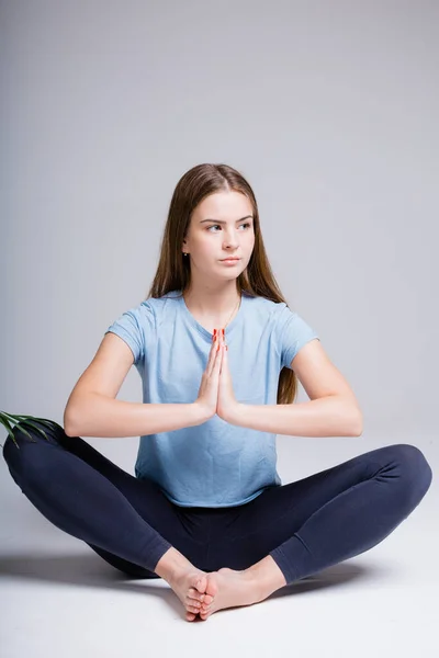 Foto vertical de uma garota europeia que se senta em uma posição de lótus com as mãos apertadas na frente de si mesma em um fundo leve no estúdio . — Fotografia de Stock