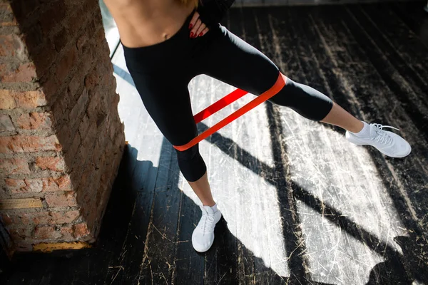 Close-up van de benen van een meisje in leggings die schudt haar binnenste dijspieren met sport rubber tape in een loft in de buurt van het raam. — Stockfoto