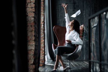 Havacı koltuğunda oturan bir iş kadınının yatay fotoğrafı ve çatı katındaki büyük bir sözleşmenin imzalanması şerefine kenarlara kağıt fırlatması..