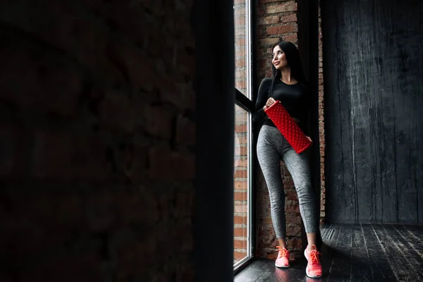 Retrato de cuerpo entero de un entrenador de fitness con polainas ajustadas apoyadas en una pared sujetando un rodillo de masaje y mirando a la ventana panorámica en el desván —  Fotos de Stock