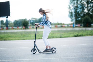 Bir kız kaldırımlı düz bir yol parkı boyunca scooter sürüyor. Açlık yürüyüşü sırasında spor aktiviteleri.