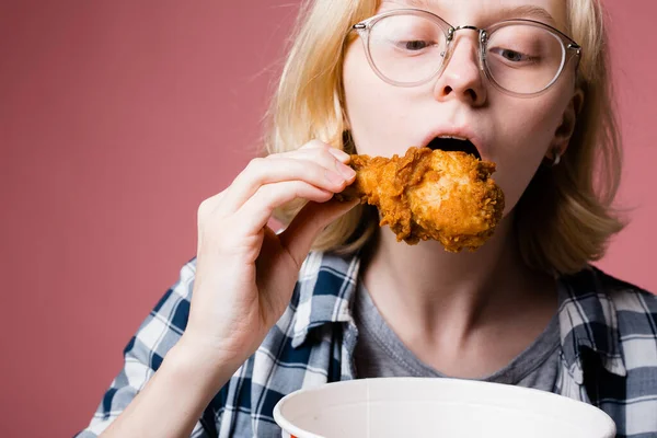 Detailní záběr blondýny dívka s brýlemi kousání lahodný kus kuřecích stehýnka v těstíčku z fast food restaurace. — Stock fotografie