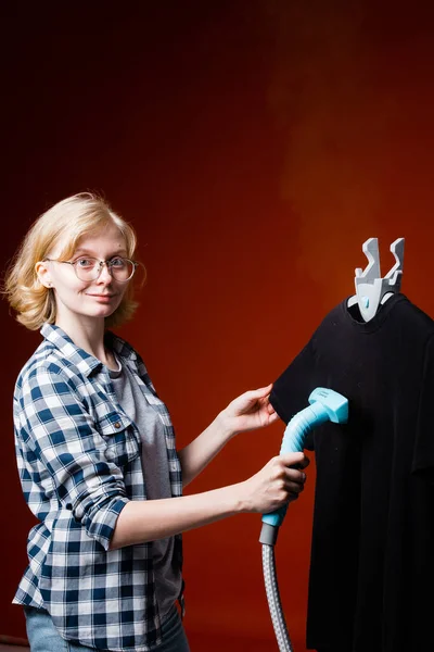 Ελκυστική νεαρή ξανθιά με γυαλιά τσαλακώνει σε μαύρο t-shirt με ένα ατμόπλοιο χέρι. Εργαζόμενος χημικός καθαρισμός για την προετοιμασία ρούχων. — Φωτογραφία Αρχείου