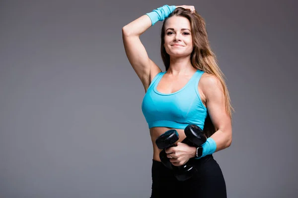 Retrato de una chica entrenadora de fitness en un top azul que tira de pesas y sonríe a la cámara sobre un fondo gris —  Fotos de Stock