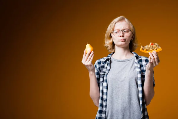 Una ragazza bionda in occhiali e una camicia a quadri tiene una pera e un pezzo di pizza tra le mani su uno sfondo arancione e guarda perplesso il cibo. — Foto Stock