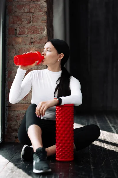 Mujer con capucha blanca que se sienta cerca de una ventana y bebe proteína de una coctelera mientras sostiene su mano sobre un cilindro de masaje rojo para estudiar la fascia muscular . —  Fotos de Stock