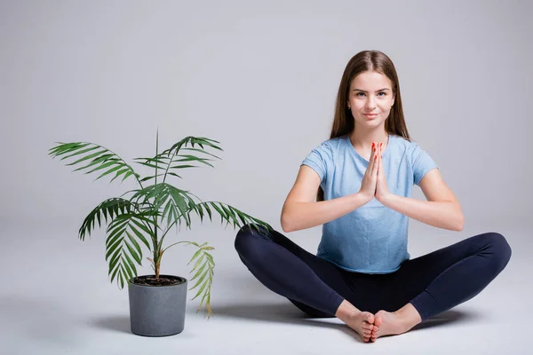 A girl yoga trainer with long hair sits in a lotus position next to a flower on a light background and meditates — Stock Photo, Image