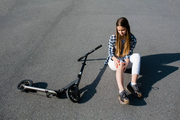 Una chica se sienta en el asfalto junto a un scooter abandonado y comprueba si tiene una contusión en la rodilla debido a una caída de un vehículo sobre el asfalto. — Foto de Stock