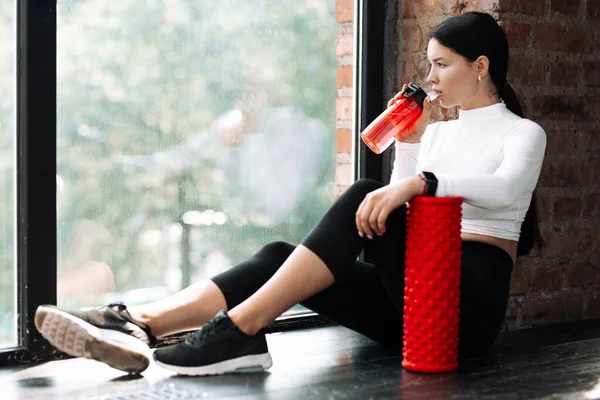 Foto de una chica asiática sentada cerca de la ventana y bebiendo agua o proteína de una coctelera roja apoyando su mano en un cilindro de masaje de espuma para fascia en el gimnasio —  Fotos de Stock