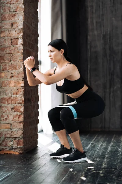 Mujer asiática en ropa deportiva negra haciendo sentadillas con expansor de muslo elástico para más resistencia en el entrenamiento de gimnasio — Foto de Stock