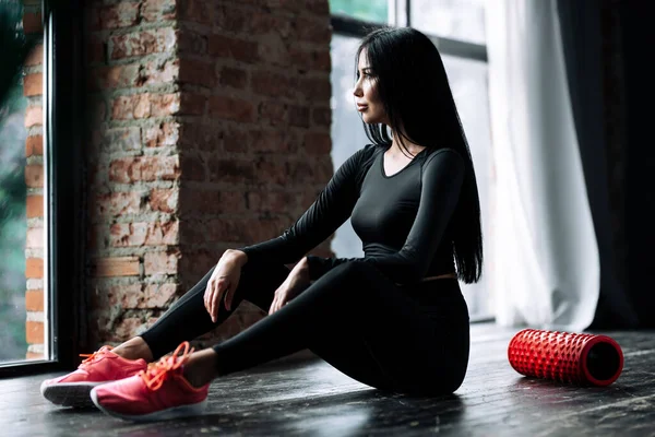 Πορτρέτο ενός κοριτσιού σε μια μαύρη αθλητική στολή και με μακριά μαλλιά κάθεται στο πάτωμα στο γυμναστήριο που massaged την περιτονία των μυών του σώματος με έναν κύλινδρο αφρού — Φωτογραφία Αρχείου