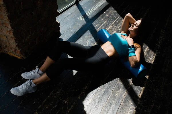 Una vista aérea de una chica de un entrenador de pilates en un top azul masajeando la fascia de los músculos de la espalda con un rodillo de espuma mientras está acostado en el suelo bajo la luz del día — Foto de Stock