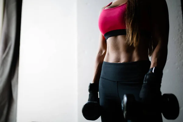 Nahaufnahme eines durchtrainierten Oberkörpers eines Kickboxermädchens in einem rosafarbenen Oberteil, das zum Training schwere Hanteln in den gesenkten Händen hält — Stockfoto