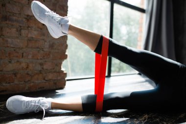 Pencerenin yakınındaki bir tavan arasında yerde elastik bir genişletici kullanarak bacak kaslarını çalıştıran bir kadının tozluklarıyla yakın plan çekimleri.