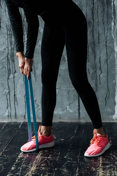 Una mujer en un chándal negro haciendo curvas con tirar hacia atrás un expansor de goma elástica usado en su pierna para entrenar los músculos de los brazos y las piernas —  Fotos de Stock