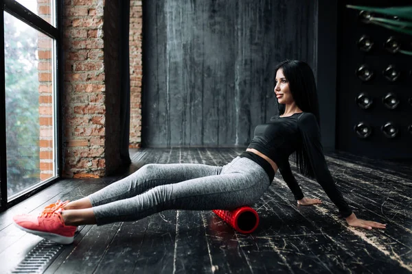 Ung kvinna pilates tränare i sportkläder tränar fascia i ryggmusklerna i benen medan du sitter på ett skum massage cylinder framför fönstret — Stockfoto