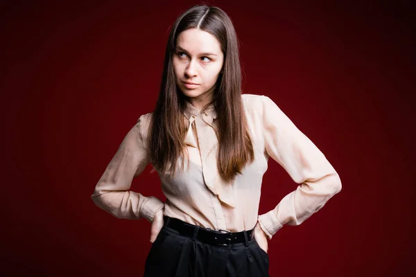 Μια μελαχρινή με ελαφρύ πουκάμισο κρατά τα χέρια της στη ζώνη της και κοιτάζει στο πλάι. Νεαρή γυναίκα με ρούχα γραφείου σε κόκκινο φόντο. — Φωτογραφία Αρχείου