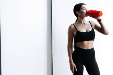 Asyalı genç bir kadının portresi beyaz arka planda siyah kaplamalı protein ya da sudan su içiyor.