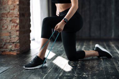 Genç bir kadın dizinin üstünde duruyor ve bir çatı katında kollarının kaslarını şişirmek için bacağıyla elastik bir genişleticiyi eziyor.