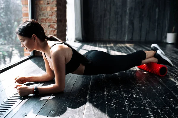 Vista lateral de una mujer joven apoyada en sus manos en el suelo para masajear la fascia de los músculos de las piernas en un desván —  Fotos de Stock