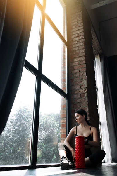 Uma visão distante de uma jovem mulher sentada perto de uma janela panorâmica e relaxante depois de exercitar a fáscia dos músculos do corpo — Fotografia de Stock