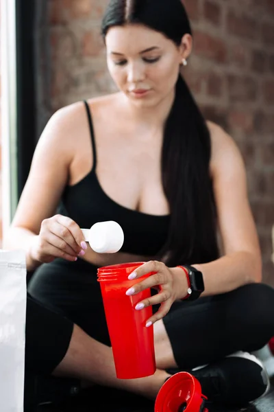 Young woman trainer in a black top with a spoon puts protein in a shaker during a break from workout sitting next to the window in the gym