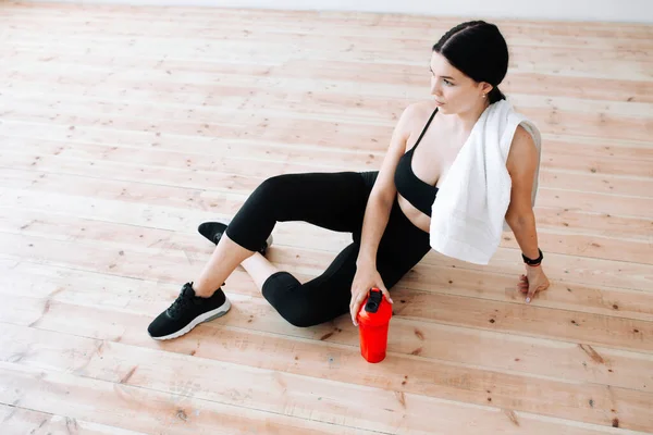 Молода брюнетка в чорному костюмі сидить на дерев'яній підлозі з білим рушником на плечі і спортивною пляшкою для тренувань після ранкового тренування . — стокове фото