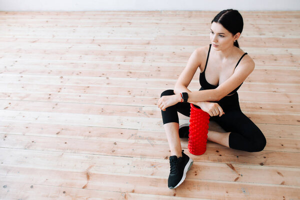 A brunette Asian woman in sportswear sits on the floor, leaning on a massager for muscle fascia and getting ready for a workout.