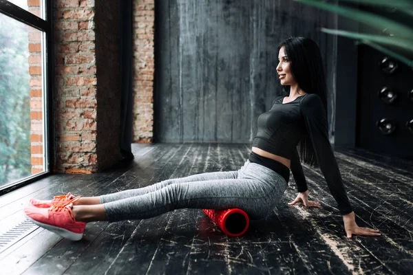 Una atleta asiática durante una clase de yoga colocó un masajeador de rodillos para calentar los músculos de la fascia debajo de sus caderas y sostiene sus piernas del suelo . — Foto de Stock