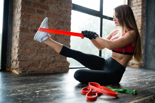 Vista lateral de una mujer joven con el pelo largo en una parte superior rosa que pone en un expansor de goma elástica en sus piernas para entrenar los músculos de las piernas —  Fotos de Stock