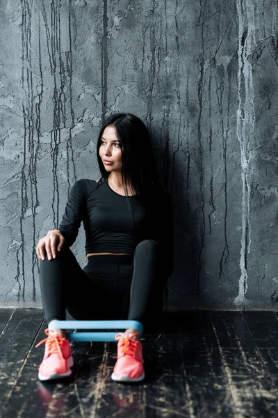Joven entrenadora de pilates en ropa negra y ajustada, se sienta en el suelo en un loft con un expansor de goma deportiva en sus pies . —  Fotos de Stock