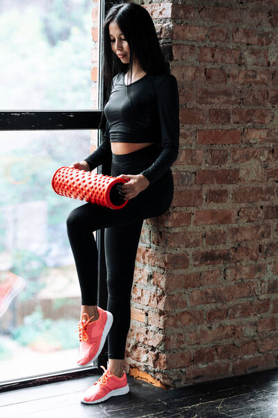 Young woman in a black tight-fitting sportswear who stands near a panoramic window and mashes a quadriceps fascia with a massage cylinder.