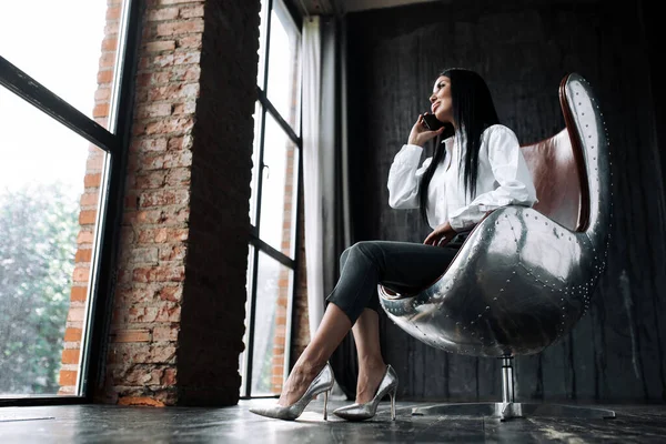 Bottom view of a beautiful woman with long hair sitting in leather chair in a loft and talking on the phone looking out the panoramic window — Stock Photo, Image