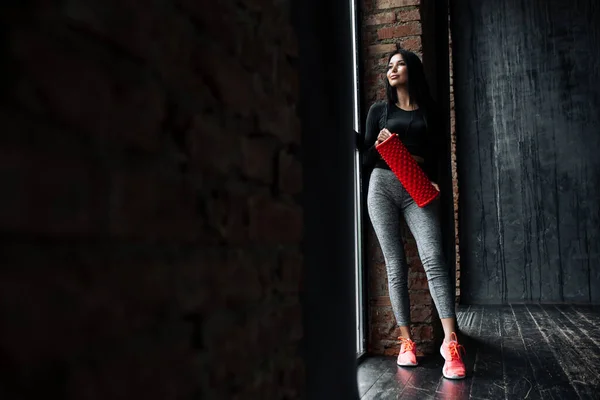 Mujer joven india en ropa deportiva está descansando después del final de una sesión de entrenamiento intenso y estirando los músculos con un rodillo deportivo rojo con un masajeador . —  Fotos de Stock